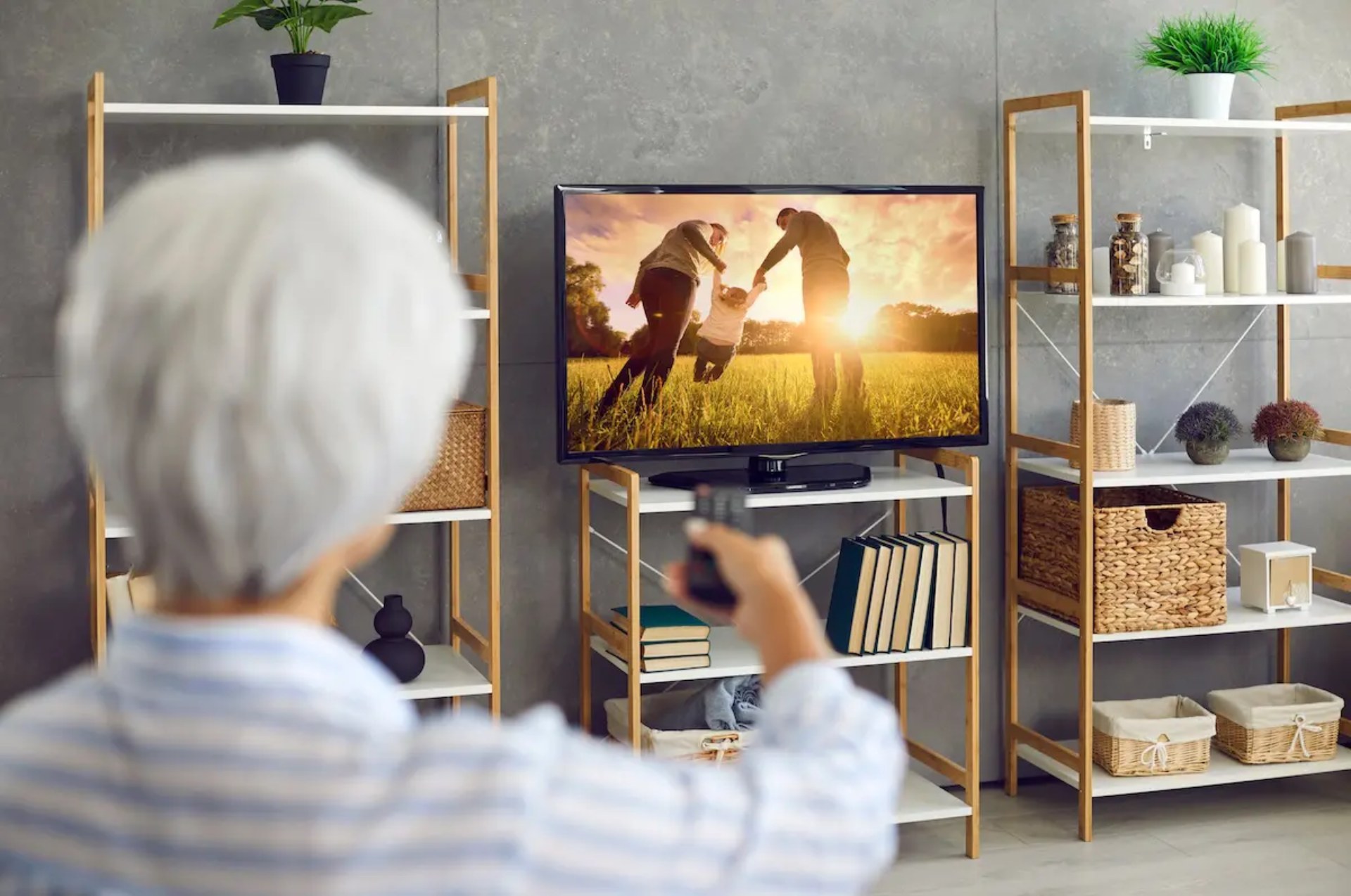 A senior person holding a remote watches a television showing a family playing outdoors, set in a cozy room with shelves and decor.