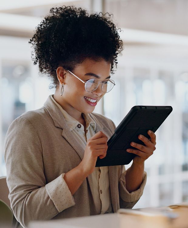 A woman wearing glasses and a blazer uses a digital tablet in a bright office setting.