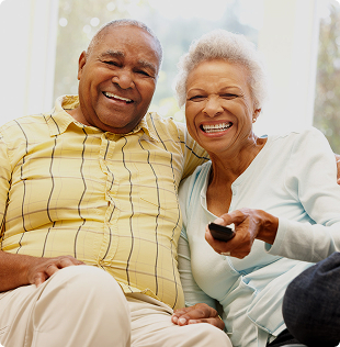 Two senior residents sitting together on a couch smiling, one holding a television remote in a bright living room setting.
