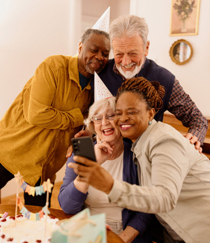 Four elderly residents wearing party hats smiling and taking a selfie around a table with a birthday cake.