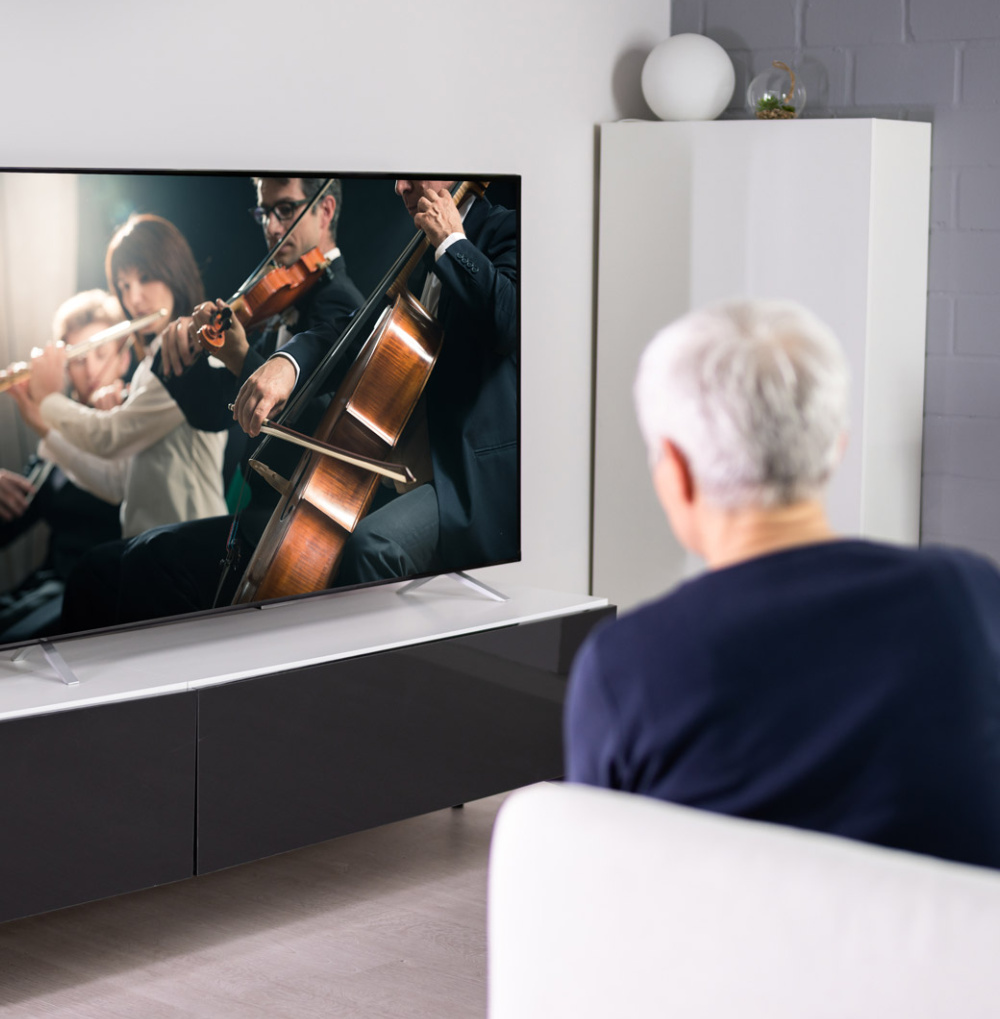 A senior watches a classical music performance with violin, cello, and flute on television in a living room setting.