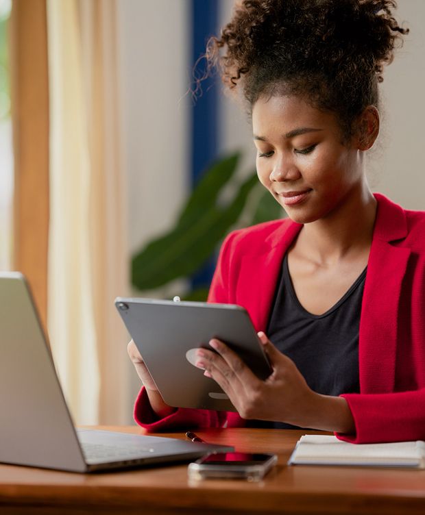 Person using a tablet at a table with a laptop, smartphone, and notebook in a well-lit indoor area.