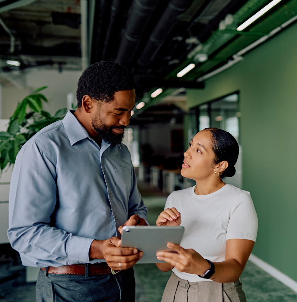 Two professionals discussing work while looking at a tablet in an office with green walls and plants.