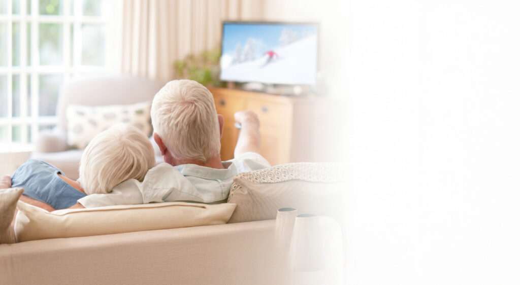 Two older adults sitting on a couch watching a skiing program on television in a cozy living room setting.