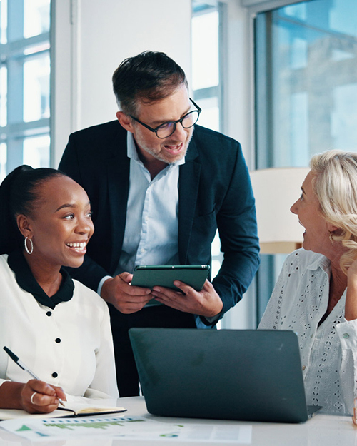 Three professionals collaborating in an office setting with a laptop and tablet, engaged in discussion around a table.