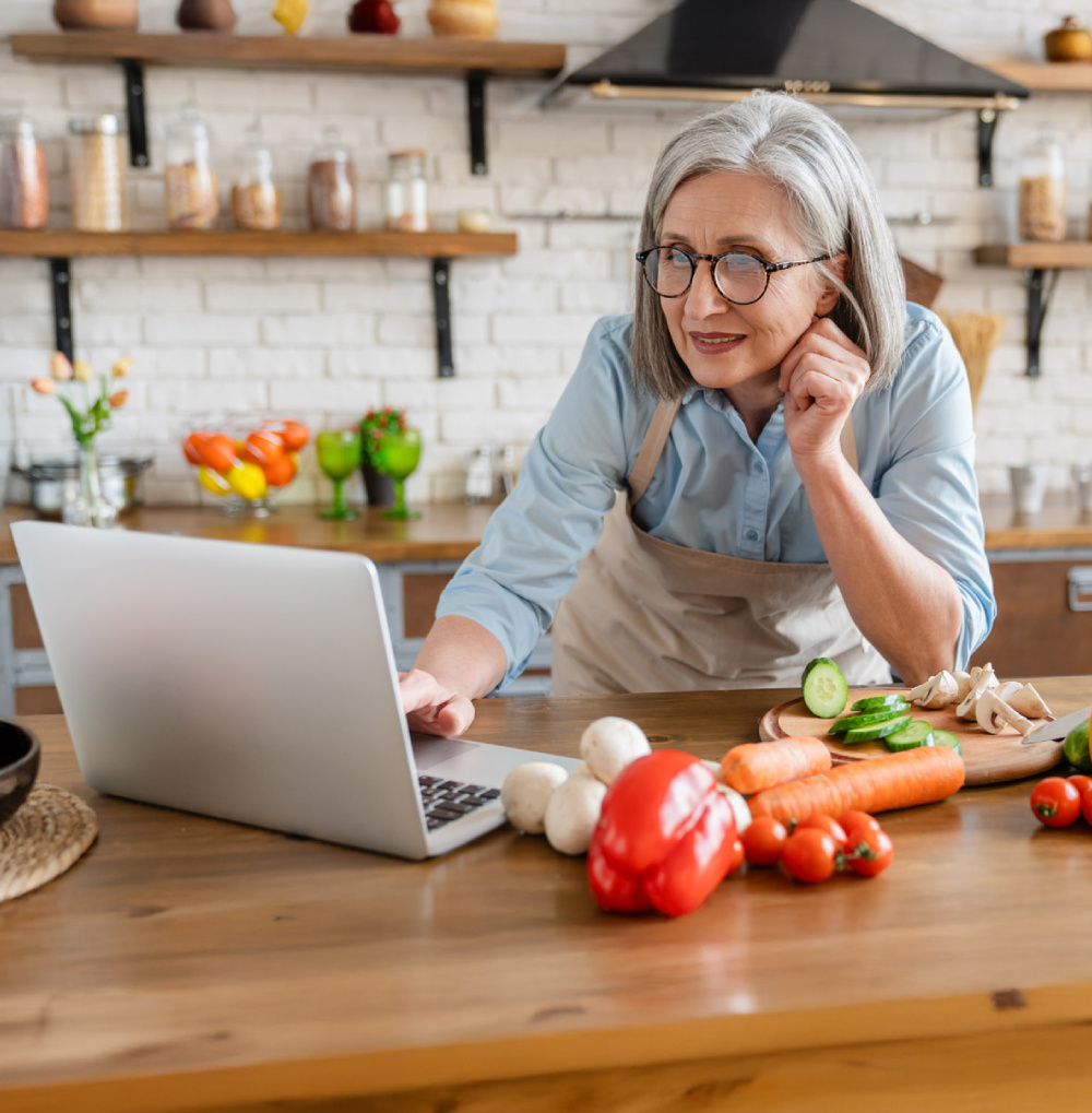 Older woman in an apron preparing vegetables in a kitchen while using a laptop on a wooden table.