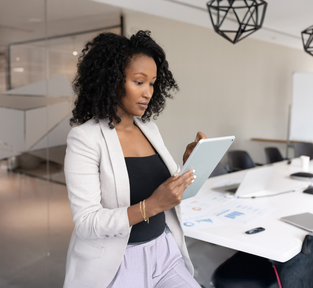 A professional woman stands in an office using a tablet near a work table with papers and a laptop.