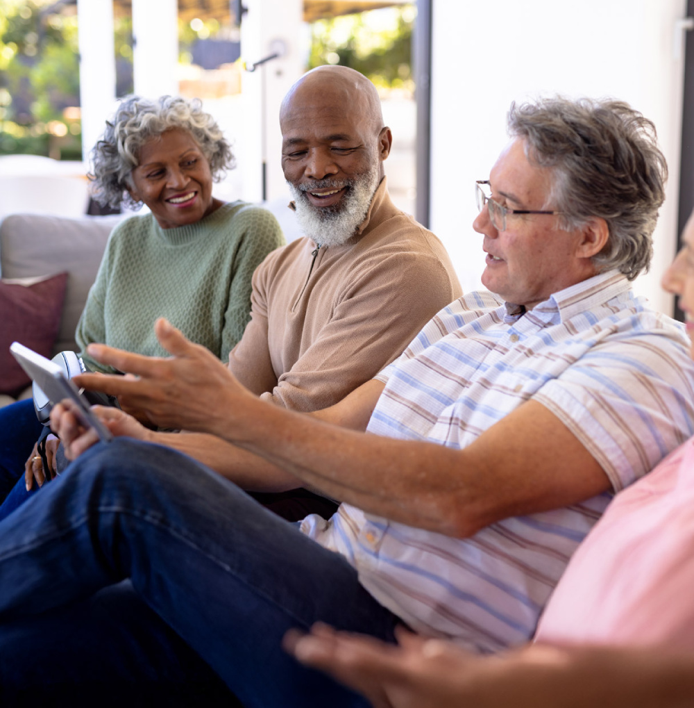 Group of seniors sitting on a couch and engaging in a conversation while sharing a tablet indoors.