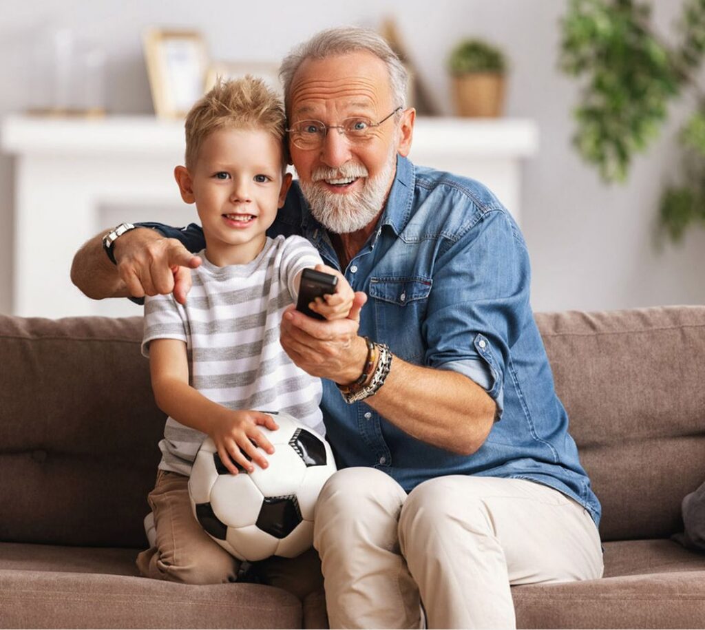 Elderly man and young boy sitting on a couch smiling with a soccer ball and a remote control in a living room.