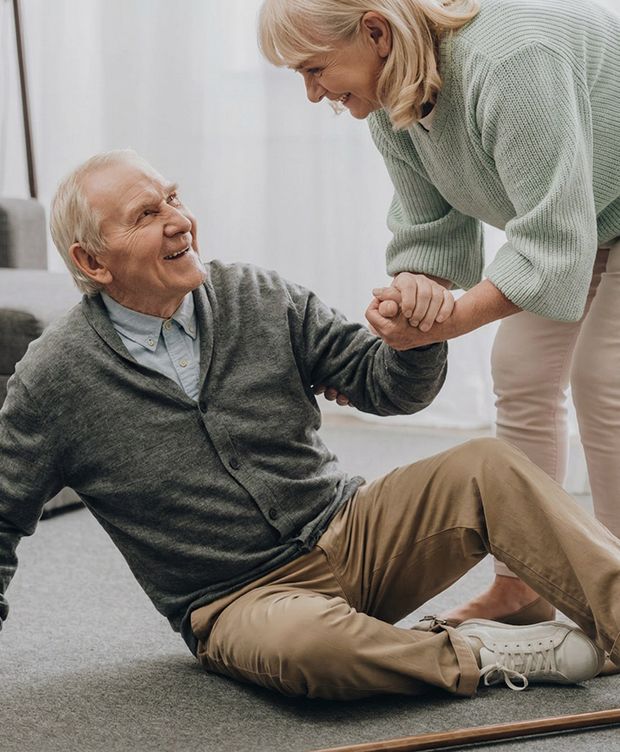 An older man sitting on the floor is helped up by a smiling older woman holding his hands in a warm home setting.