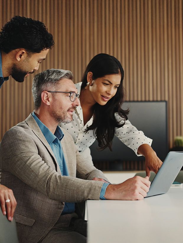 Three adults collaborating around a laptop in a modern office setting with wooden panel walls.