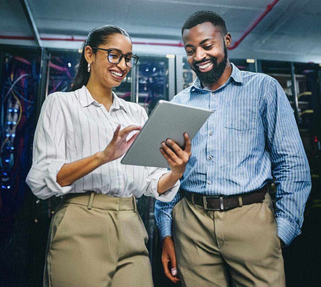 Two professionals standing and smiling while viewing a tablet in a room with servers.