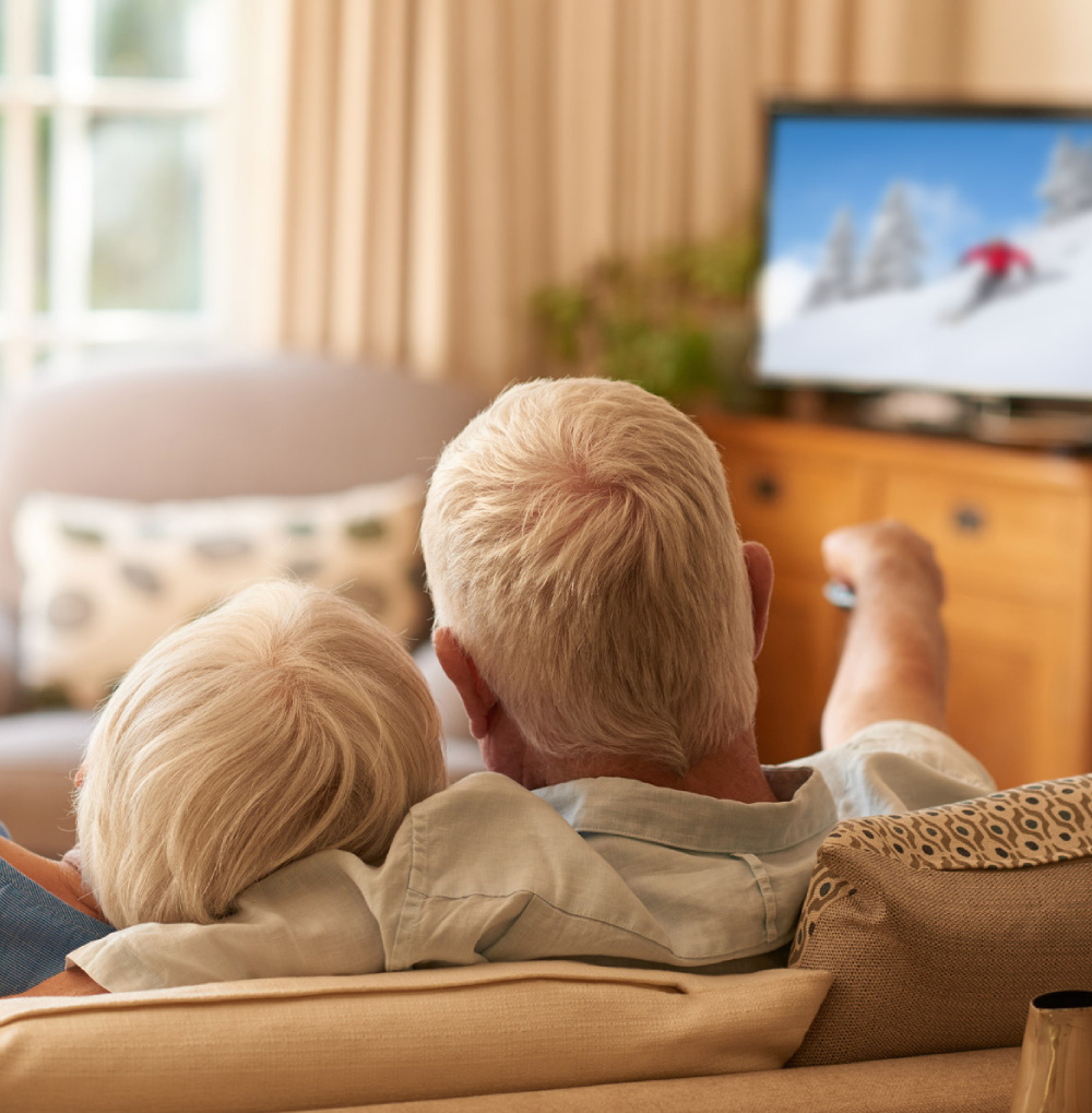Two elderly individuals sitting closely on a couch watching a winter sports program on television in a comfortable living room.