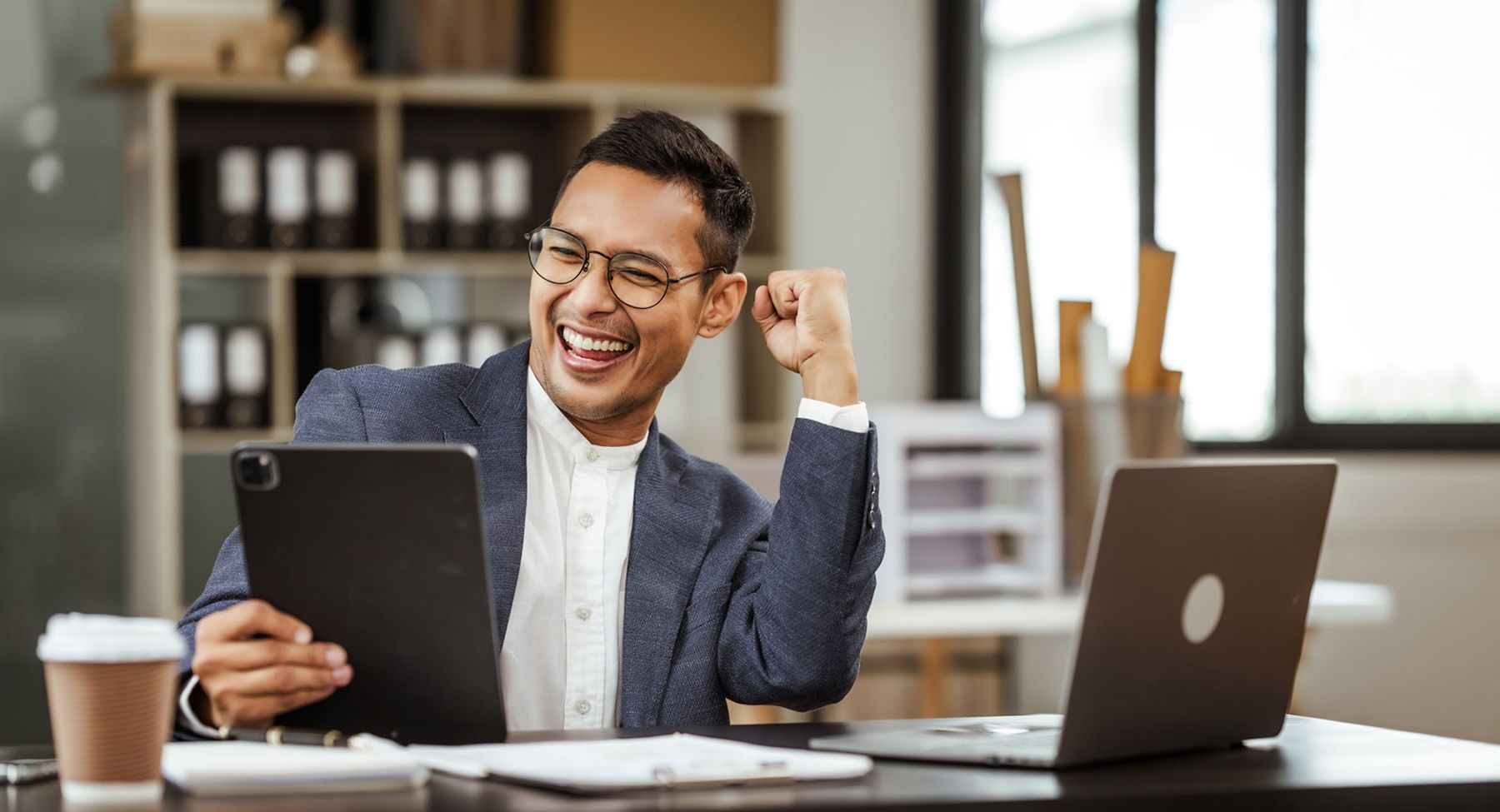 Man at office desk holding a tablet and expressing joy with a fist pump near laptop and coffee cup.