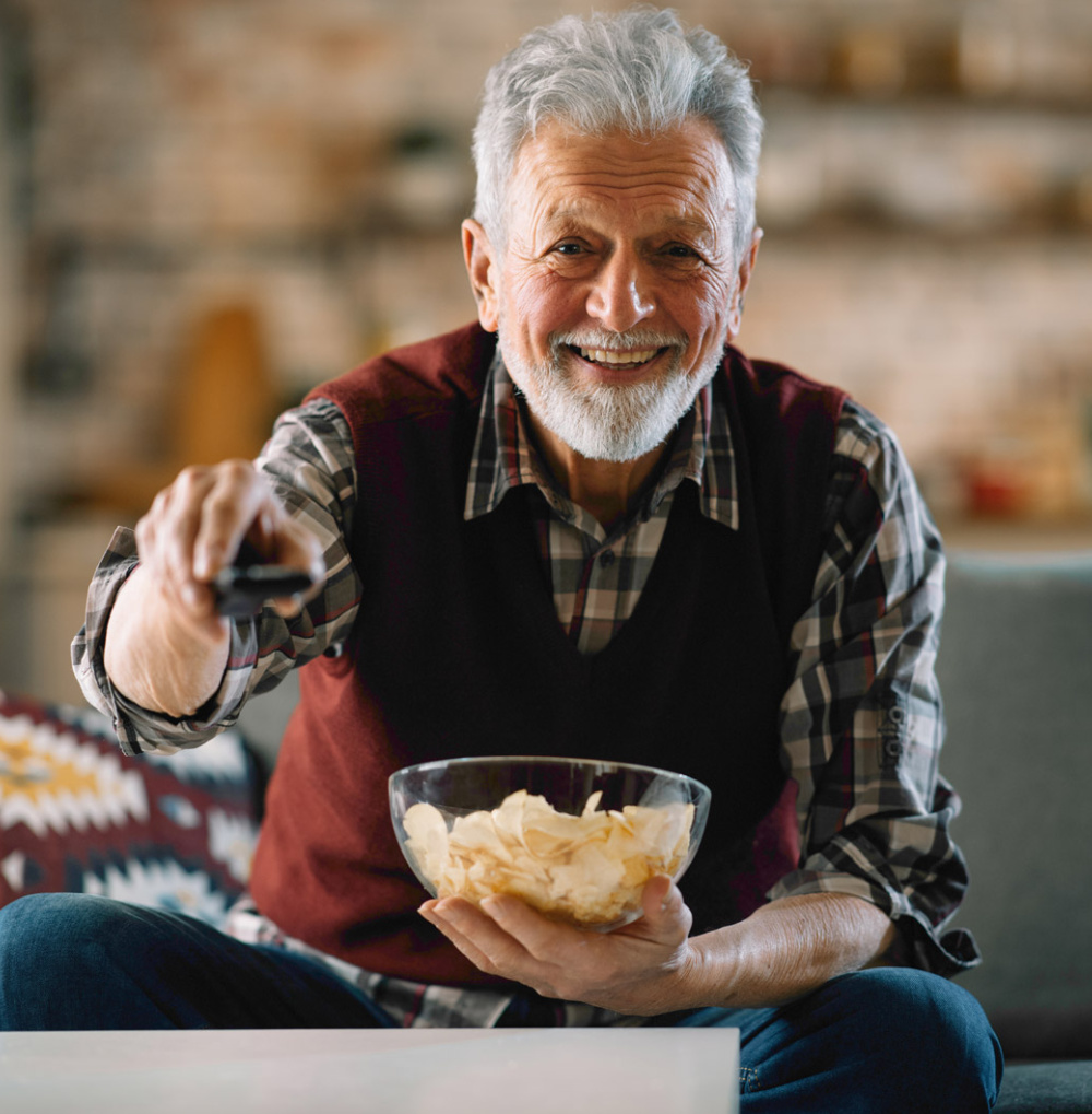 Smiling older man holding a bowl of chips and a remote control while sitting on a couch.