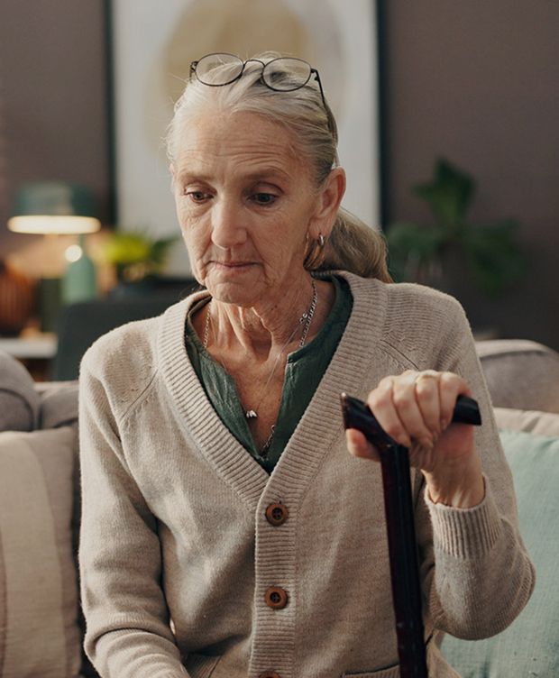 Older woman sitting on a couch indoors, holding a walking cane and wearing glasses on her head.
