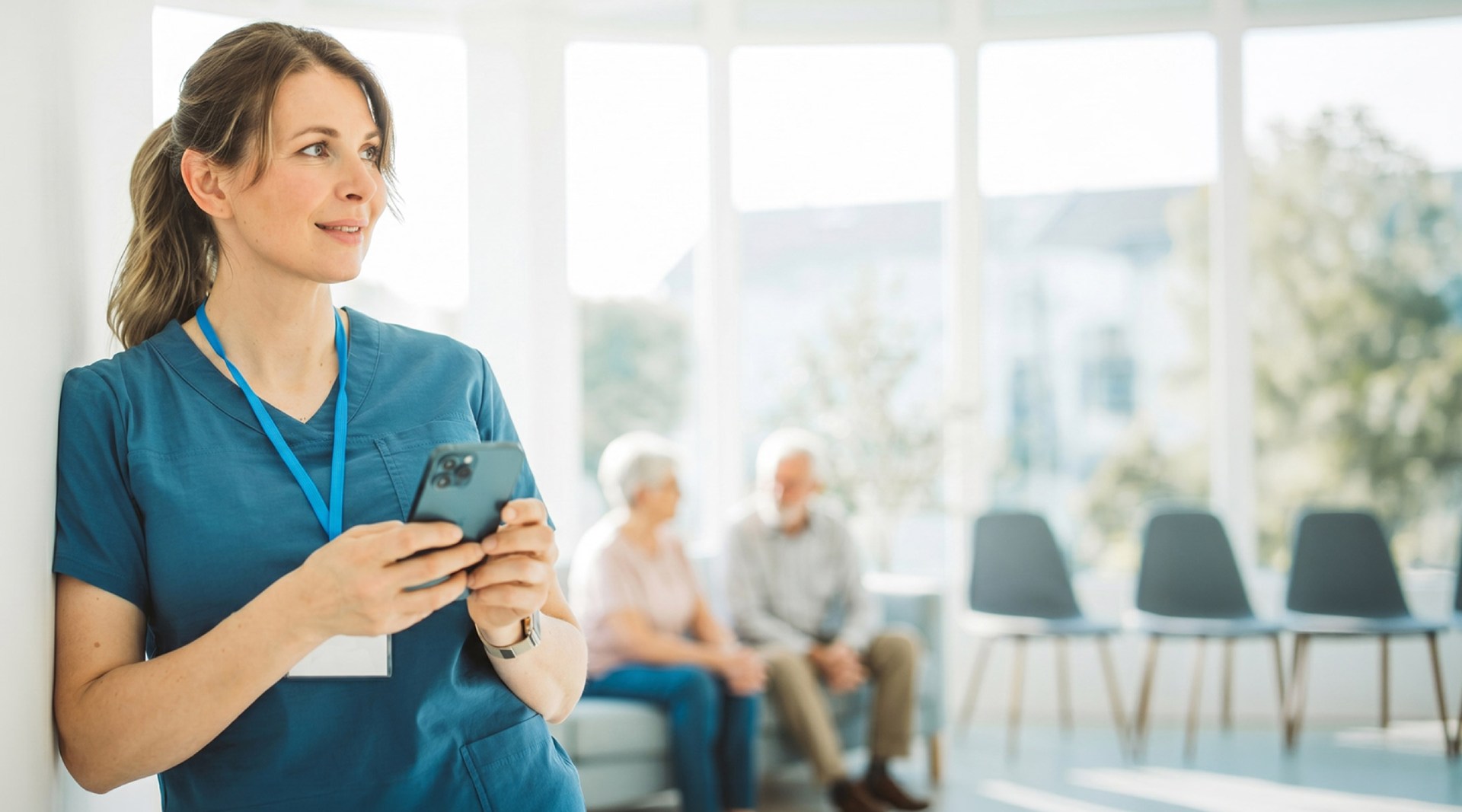 Caregiver in blue scrubs holding a smartphone in a bright senior living lounge with two seated elderly residents in the background.