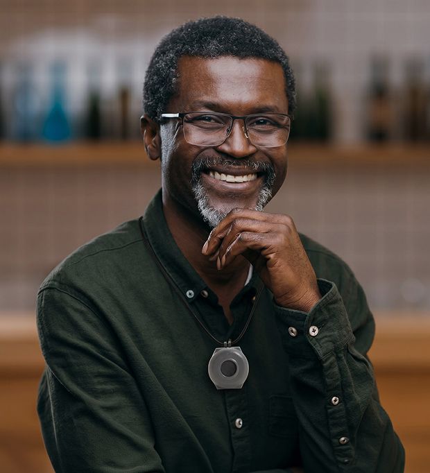 Man smiling indoors with glasses and pendant necklace, posing with hand near chin.