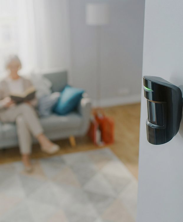 Motion sensor installed on wall in a living room with a person reading on a couch in the background.