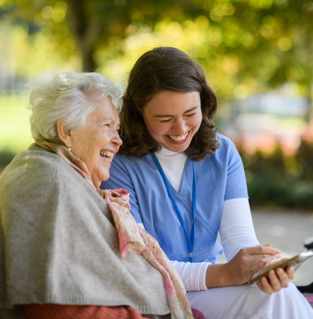 Elderly woman and caregiver sitting outdoors laughing together while looking at a smartphone.