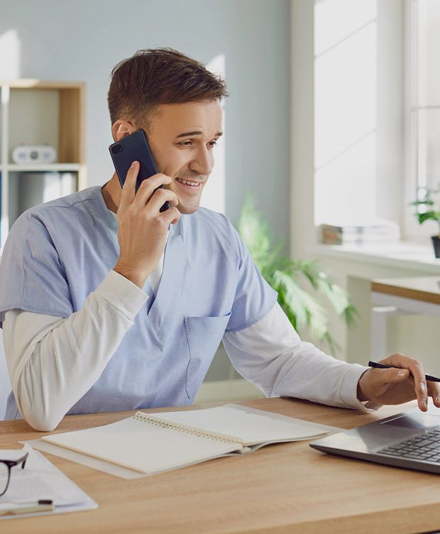 A care worker in scrubs talking on a smartphone and using a laptop at a desk in a bright office environment.