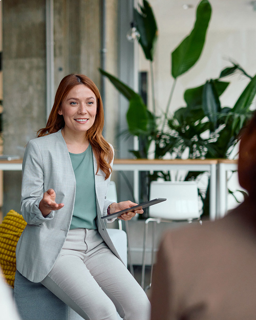 Woman holding a tablet and engaging in discussion with another person in a modern office setting with plants.