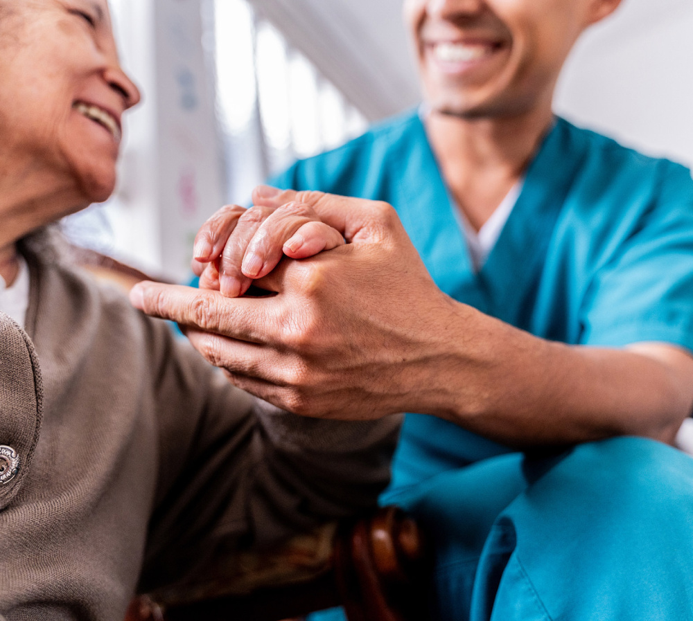 Caregiver in blue scrubs holding hands with a smiling elderly resident, conveying warmth and support in a senior living setting.