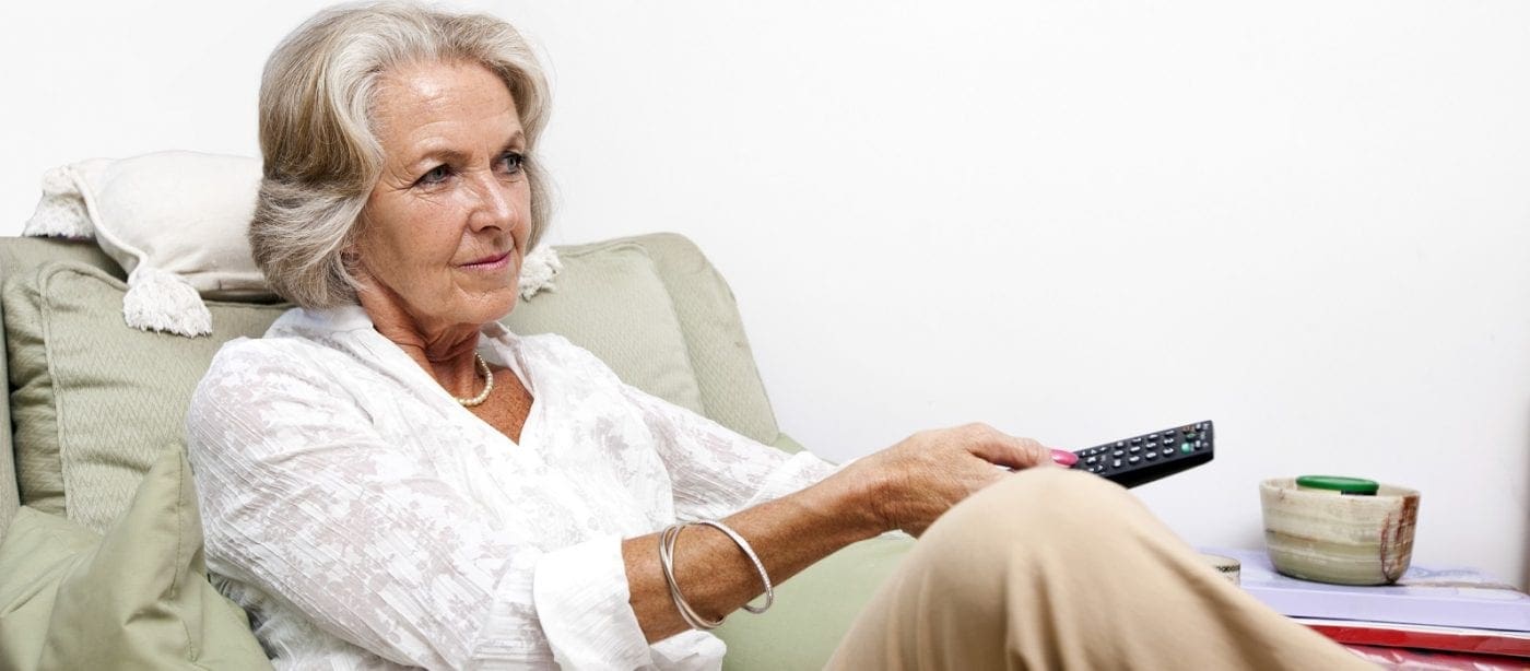 A resident seated on a couch holding a TV remote watching television in a comfortable living room setting.