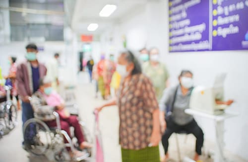 Residents and staff in a care facility corridor with wheelchairs and signage, some wearing face masks.