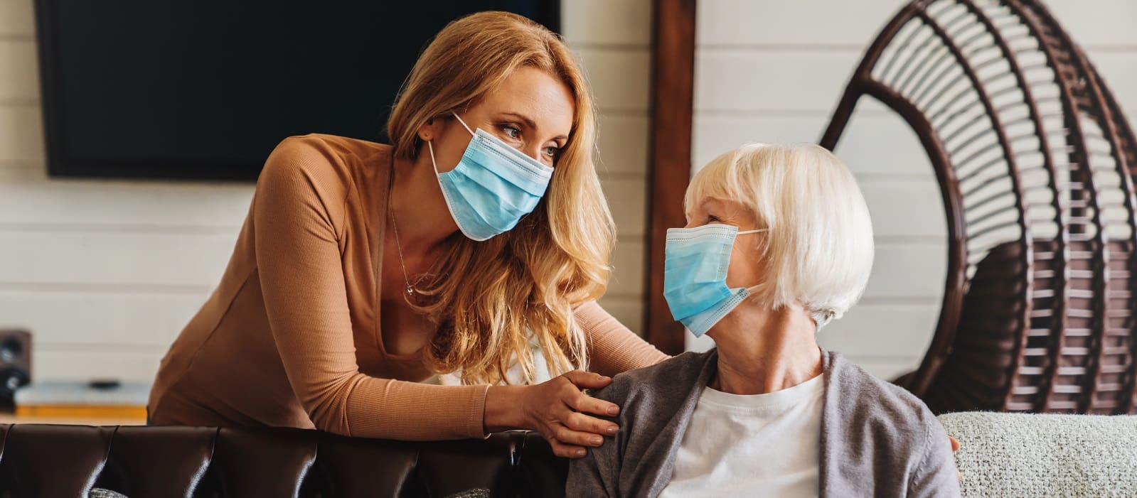 Younger woman wearing a face mask leaning over and gently supporting an older masked woman seated indoors