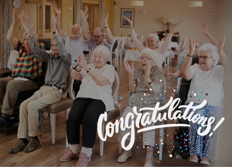 Senior residents seated in a community room clapping and raising hands during a social activity.