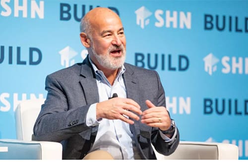 Man in a suit jacket speaking and gesturing during a corporate panel discussion with SHN and BUILD backdrop.