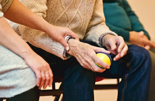 Two seated individuals engaged in hand exercise using a small ball, showing supportive interaction in a senior living environment.