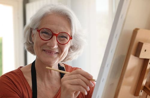 An elderly resident painting on a canvas at an easel in a bright room.