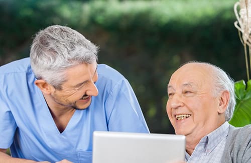 A healthcare professional and an older adult smiling and engaging with a tablet outdoors in a senior living community setting.