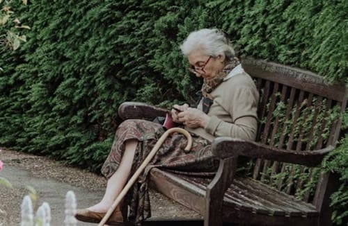 Resident sitting on a wooden bench in a garden using a smartphone with a cane resting nearby and greenery around.