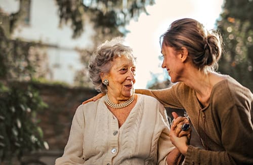 Older woman and younger woman engaged in a supportive conversation outdoors with greenery behind them.
