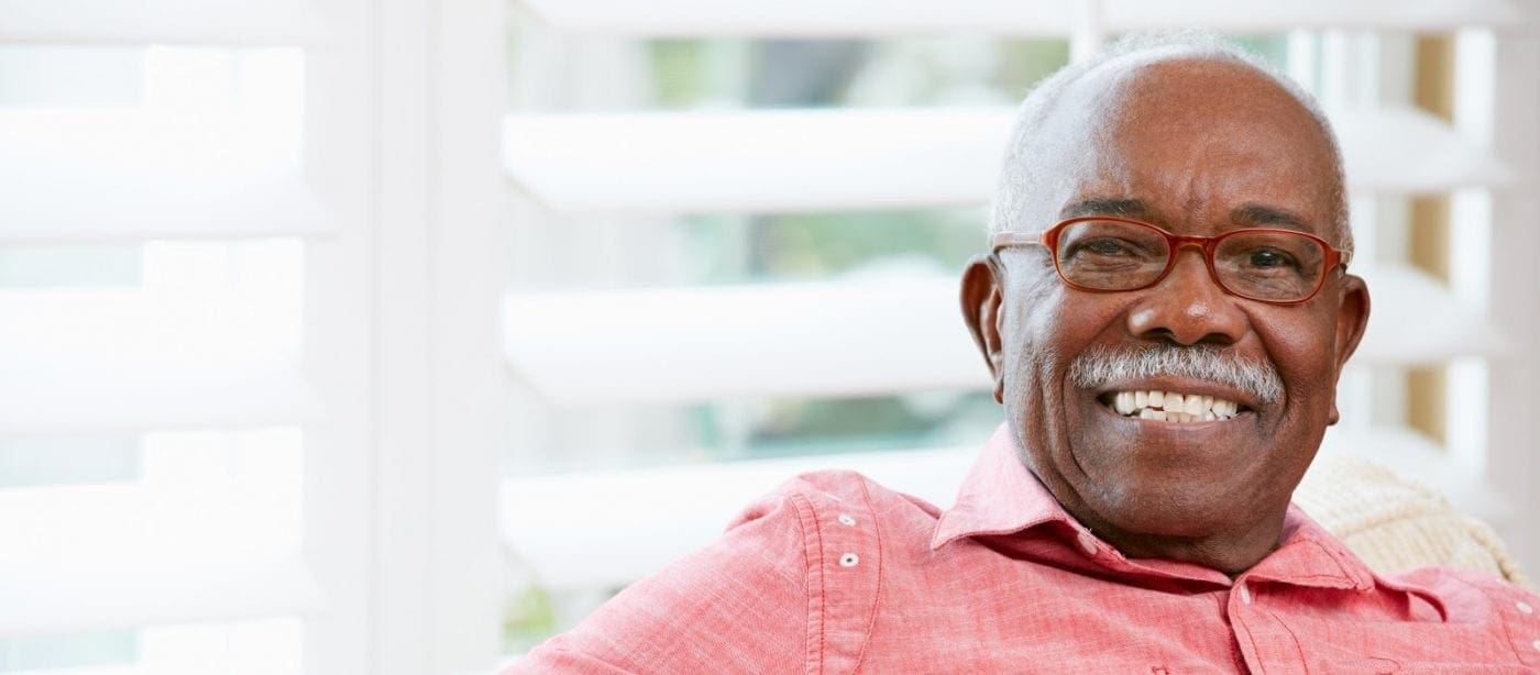 Smiling man sitting indoors near a bright window with white blinds.