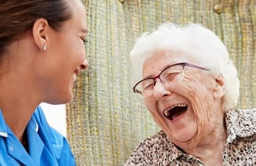 Elderly woman and caregiver sharing a joyful moment and laughing together indoors in a senior living community.