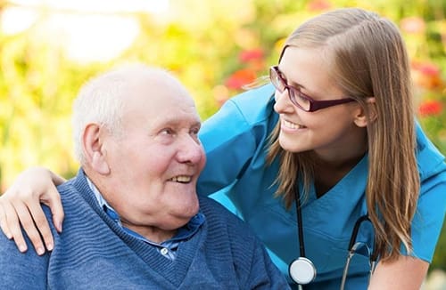 Elderly man sits outdoors smiling as female caregiver with stethoscope leans close and engages warmly.