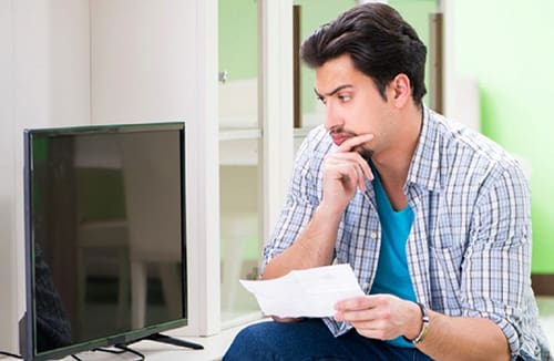 A man sitting indoors looking thoughtfully at papers near a television in a community living area.