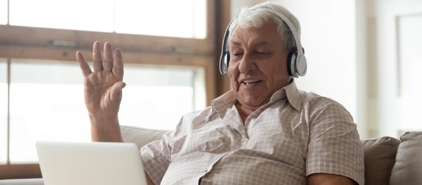 An elderly man wearing headphones sitting on a couch waving during a video call on a laptop in a comfortable indoor setting.