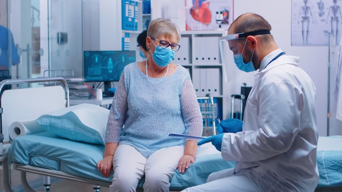 Senior woman wearing a mask sitting on a medical bed while a healthcare professional with protective gear discusses notes in a clinical room.