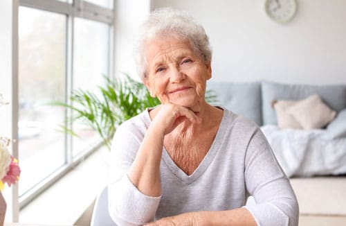Elderly woman sitting indoors by a bright window with a plant and sofa in the background.
