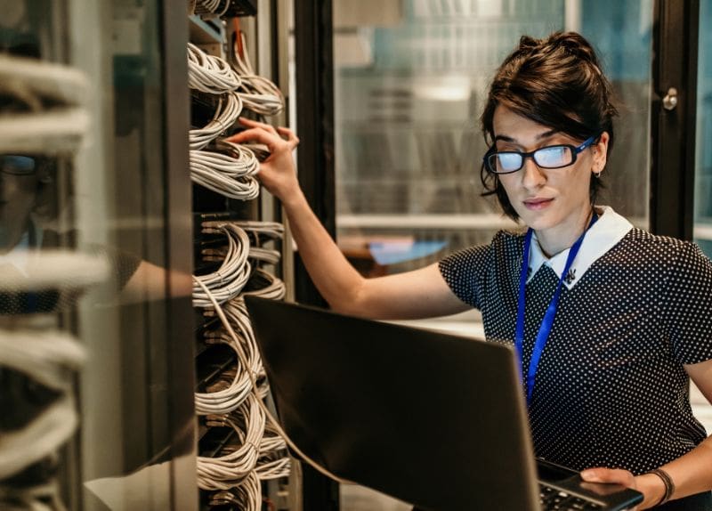 A woman works with network cables and a laptop in a server room environment.