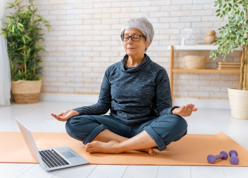 Older woman sitting cross-legged on a yoga mat indoors meditating with closed eyes near a laptop and dumbbells.