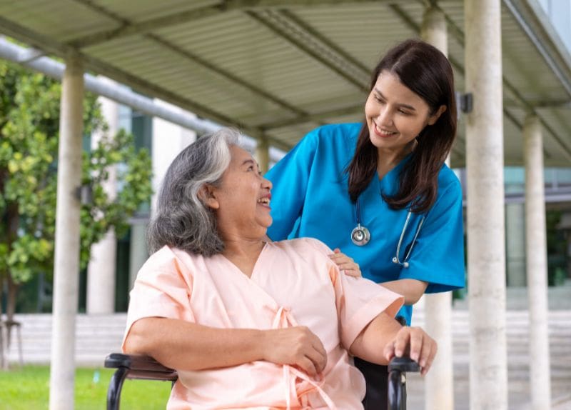 A senior resident in a wheelchair outdoors smiling and interacting with a healthcare professional in blue scrubs under a covered walkway.