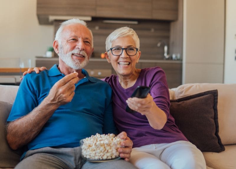 Two seniors sitting on a couch sharing popcorn and watching television together in a cozy living room.