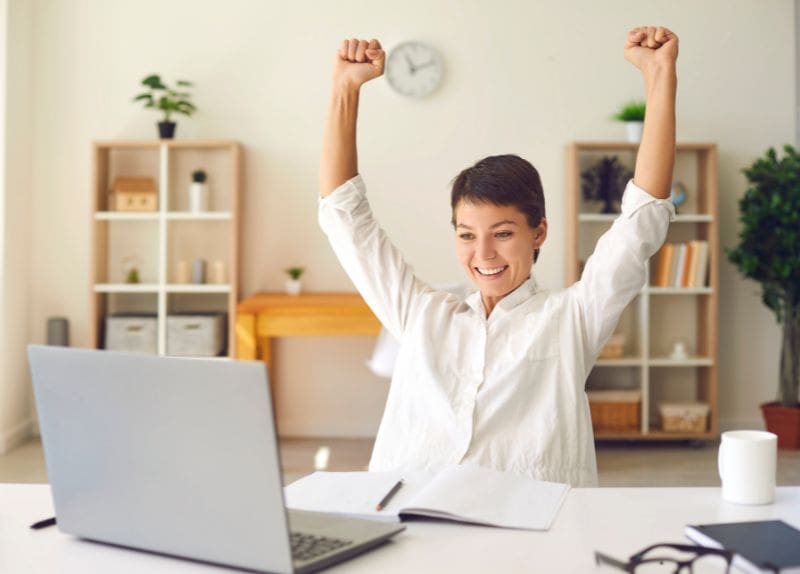 Person at desk raising arms in celebration while using a laptop in a bright room with shelves and plants.