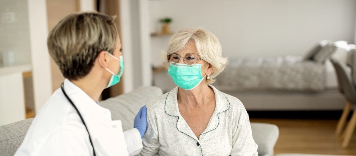 Two adults wearing face masks in a home setting during a health consultation interaction.